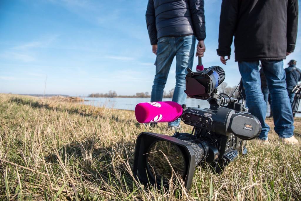 Image by Ehrecke from Pixabay. Journalists on the bank of a river with a camera on the ground.